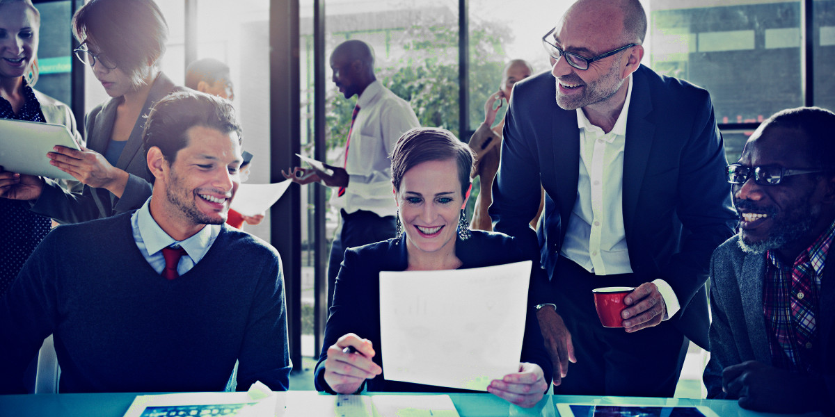 Four Smiling Team Members at Desk Looking Over Paperwork with Additional Multi-Racial Office Members in Background