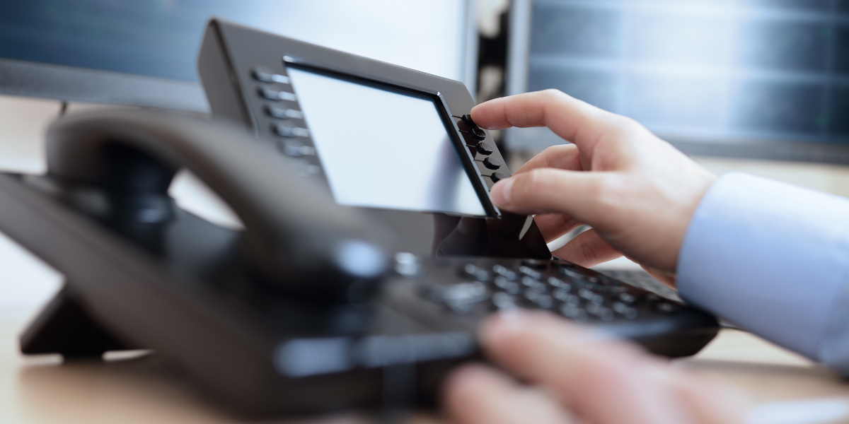 Close Up of Man's Right Hand Dialing Hold Button on VoIP Phone with Wood Desk and Computer Monitors Blurred in Background